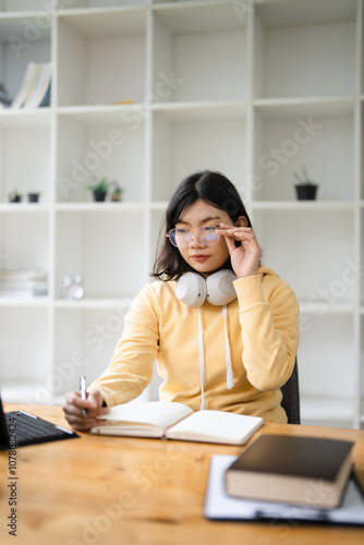 A beautiful young Asian woman in glasses and casual wear is focusing on her work on her laptop computer, reading online papers, thinking and planning her work, sitting at an outdoor table of a cafe