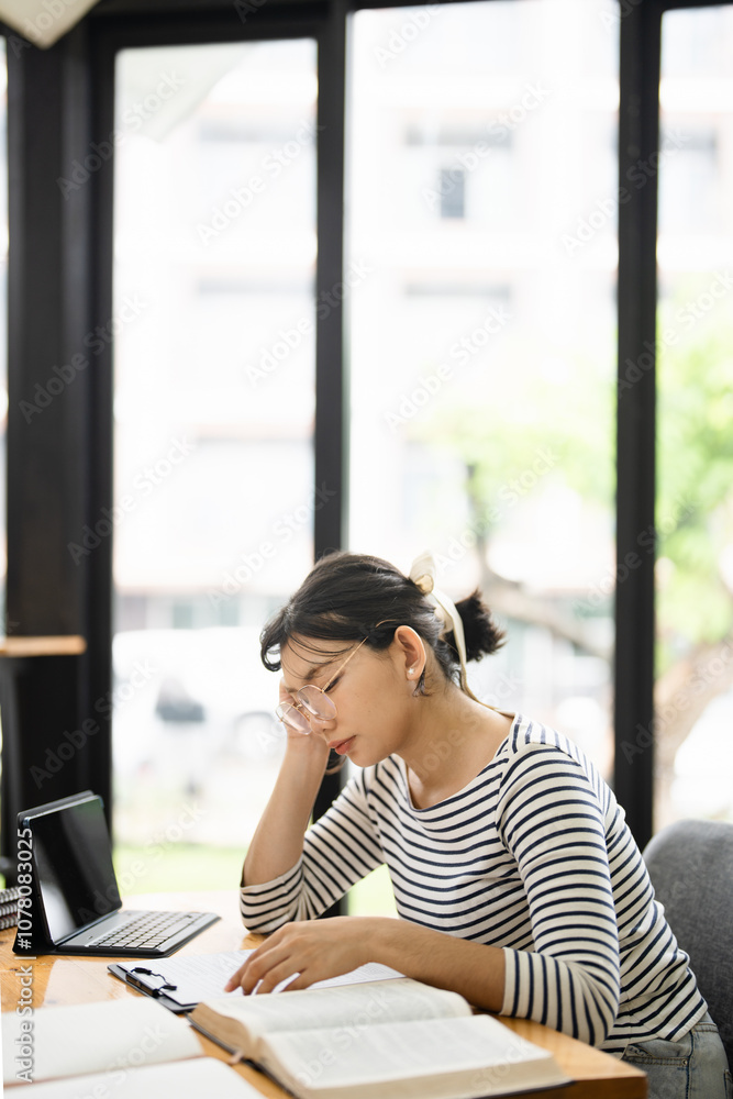 A young focused Asian woman is working remotely from a cafe, taking notes or making list in her notebook, a laptop computer on the table. A student doing homework or studying online in a coffee shop A young focused Asian woman is working remotely from a cafe, taking notes or making list in her notebook, a laptop computer on the table. A student doing homework or studying online in a coffee shop