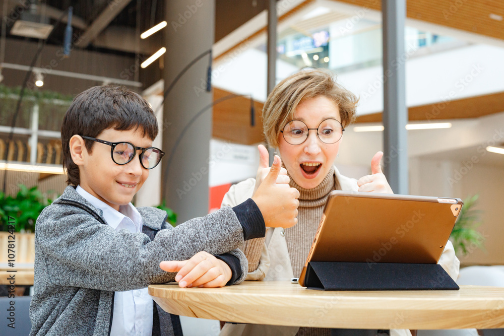 A young boy and a woman both are in glasses, looking at a tablet together, using wireless technology and communication A young boy and a woman both are in glasses, looking at a tablet together, using wireless technology and communication