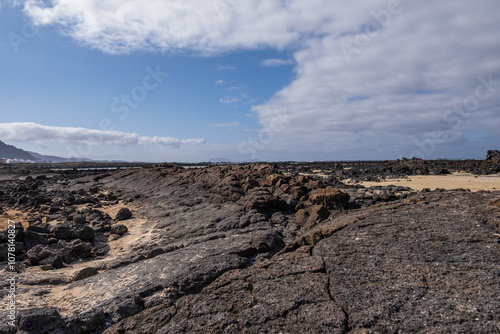 White sand beach with solidified volcanic lava flows, black rocks. Mountains in the background and sky with big white clouds. Caletón Blanco, Lanzarote, Canary Islands, Spain
