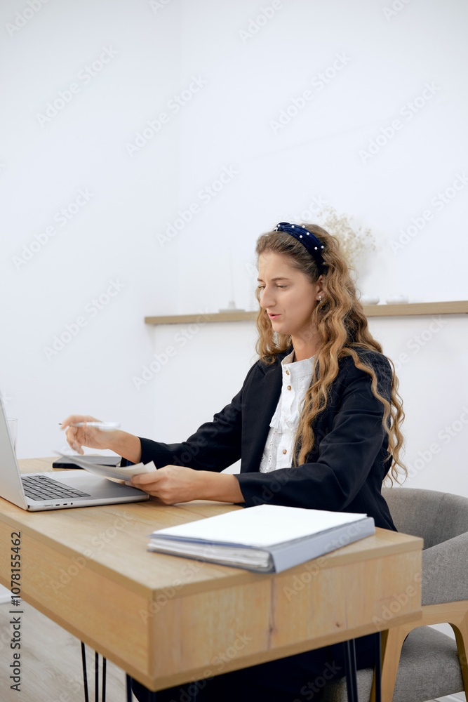 Young professional reviewing documents while working on a laptop at a modern desk Young professional reviewing documents while working on a laptop at a modern desk