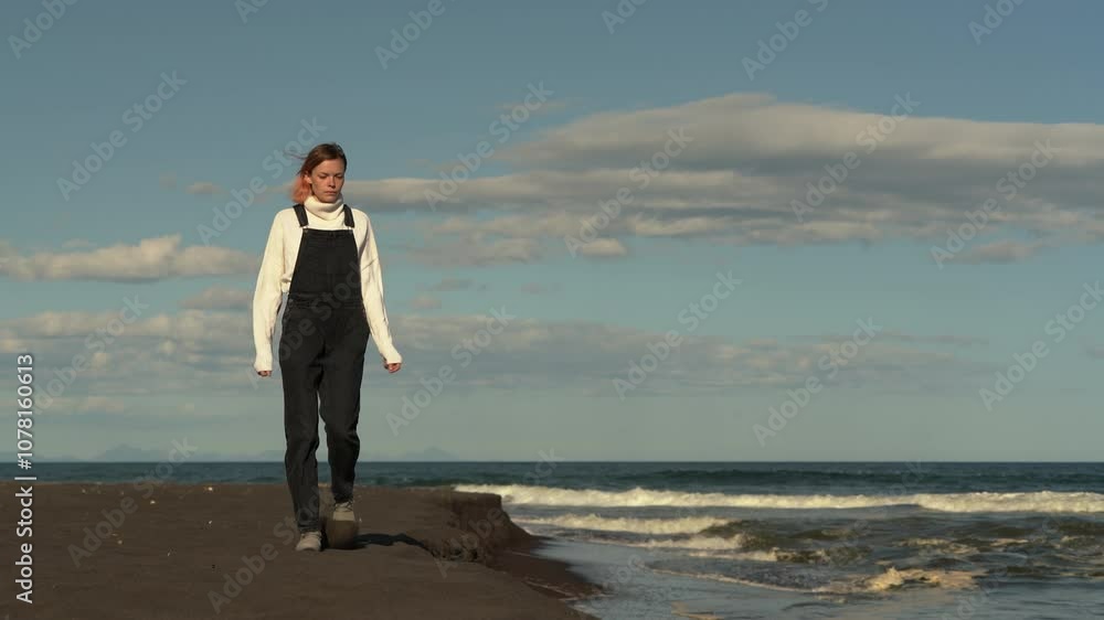 Beautiful woman is dressed in white sweater, black denim jumpsuit and trainers walking along black sandy beach during summer vacation. Real time video, cinematic handheld shot Beautiful woman is dressed in white sweater, black denim jumpsuit and trainers walking along black sandy beach during summer vacation. Real time video, cinematic handheld shot