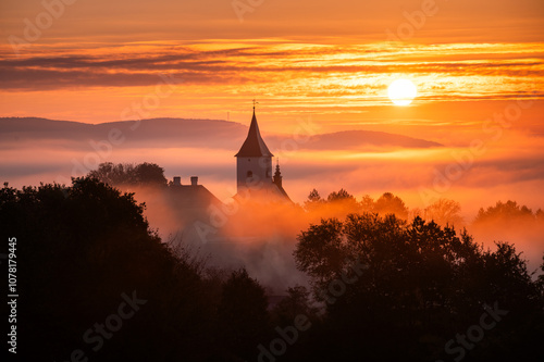 Peaceful Sunrise Over Rural Church Silhouette in Autumn, Surrounded by Mist and Glowing Orange Sky