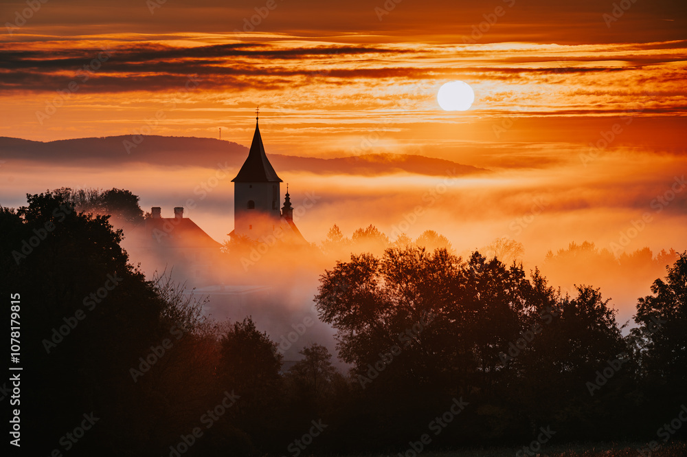 Obraz premium Silhouette of a Christian Church in a Beautiful Autumn Morning During Sunrise, Rural Landscape with Orange Sky and Soft Morning Mist Over the Village