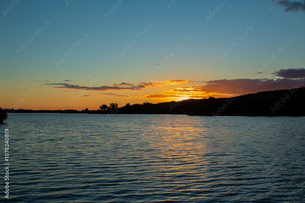 Fototapeta premium Orange colors over a river, a sunset with orange colors. The sky is tinged with warm tones, from gold to deep red, as the sun sets.