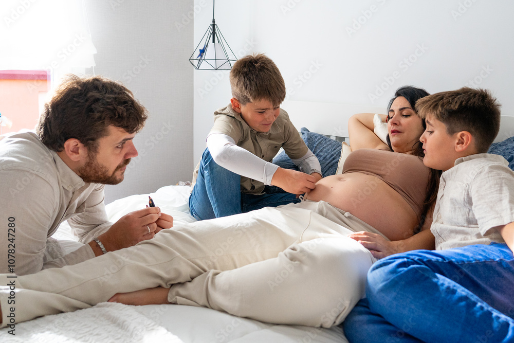 Fototapeta premium Family with pregnant mother, father and two children painting the mother's belly on the bed in the bedroom. large family