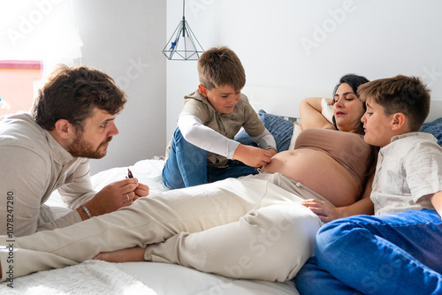 Family with pregnant mother, father and two children painting the mother's belly on the bed in the bedroom. large family