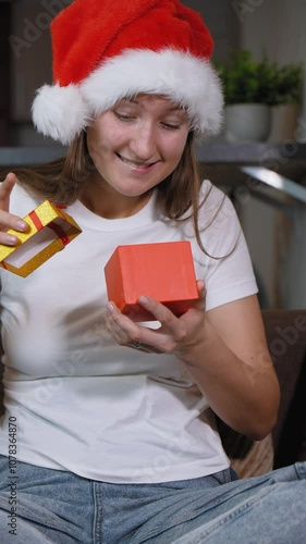 Woman in Santa hat sitting on sofa opening christmas gift. Frustration washes over her face as she opens contents inside, leading to moment of sadness, she is dissatisfied. vertical video photo