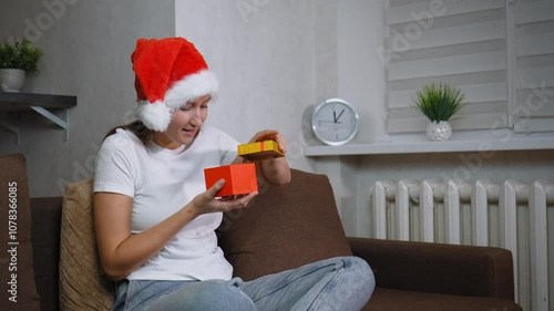 Woman in Santa hat sitting on sofa opening christmas gift. Frustration washes over her face as she opens contents inside, leading to moment of sadness, she is dissatisfied. photo