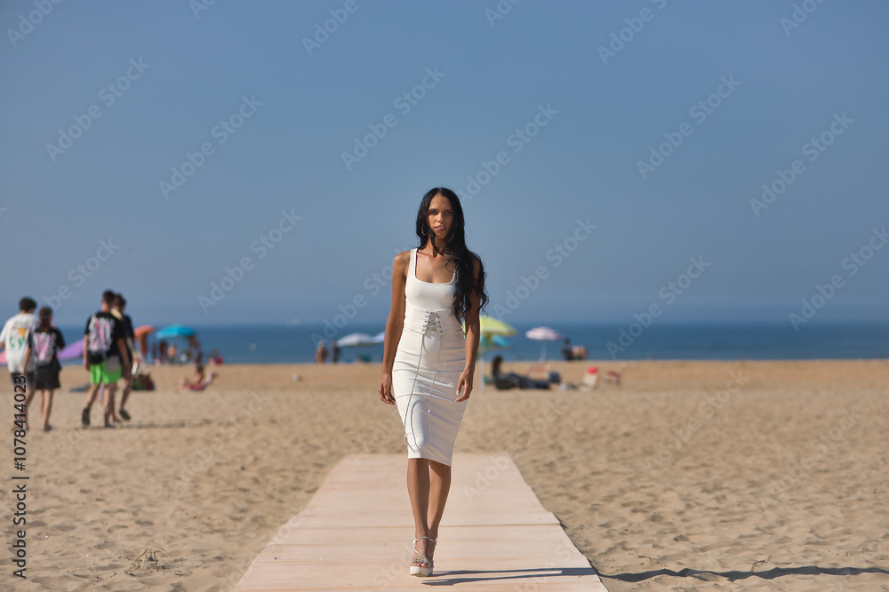 Young, beautiful, brunette, slim woman in a white dress and heels, parading as a model on a wooden walkway on the beach, with the sea in the background. Concept beauty, fashion, trendy, femininity. Young, beautiful, brunette, slim woman in a white dress and heels, parading as a model on a wooden walkway on the beach, with the sea in the background. Concept beauty, fashion, trendy, femininity.