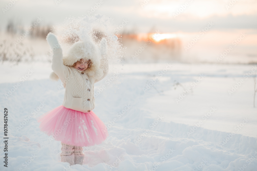 Fototapeta premium Winter lifestyle portrait of cheerful little girl in knitted sweater throwing snow in the air. Child is smiling and having fun in the snow park. Freedom and happiness concept.