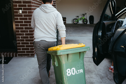 Man in a grey hoodie pushing a yellow-lidded wheelie bin photo