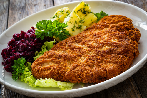 Crispy breaded fried pork chop, boiled potatoes and red cabbage served on white plate on wooden table
