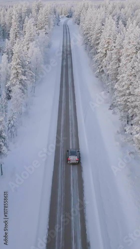 Aerial view following  a EV car driving on a road in middle of frosty forest, winter sunrise in Lapland