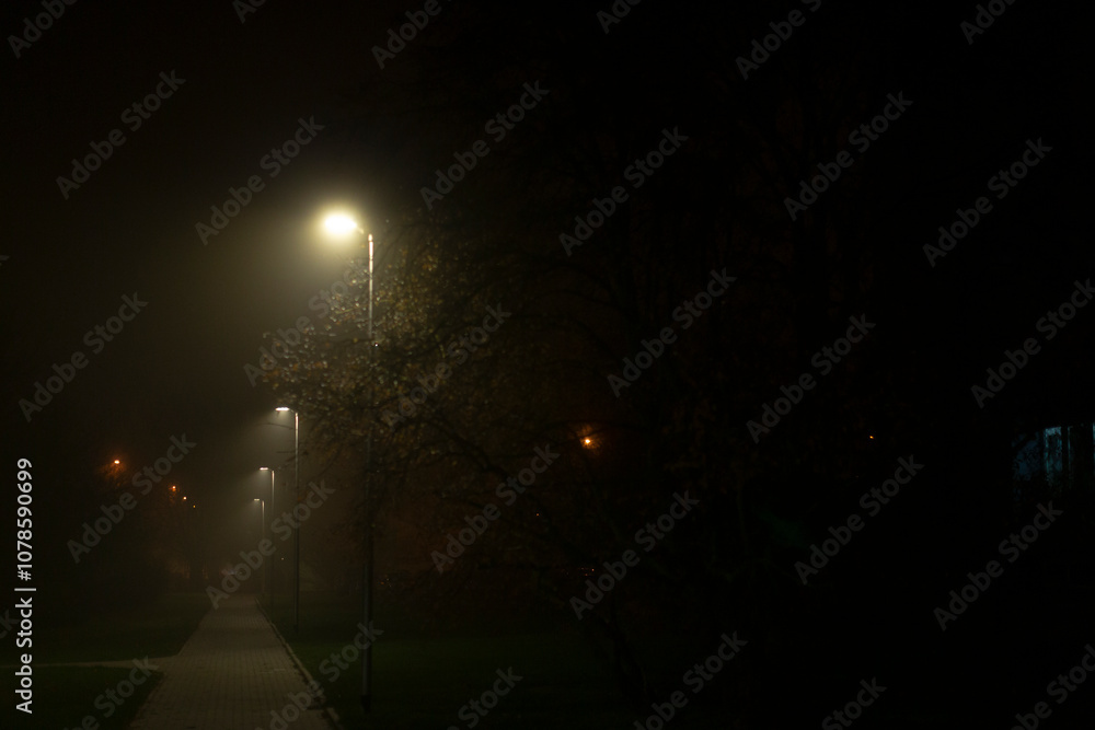 On a foggy night, a tranquil scene unfolds with a dimly lit sidewalk softly glowing under street lamps, surrounded by trees, creating a captivatingly mysterious and serene atmosphere On a foggy night, a tranquil scene unfolds with a dimly lit sidewalk softly glowing under street lamps, surrounded by trees, creating a captivatingly mysterious and serene atmosphere