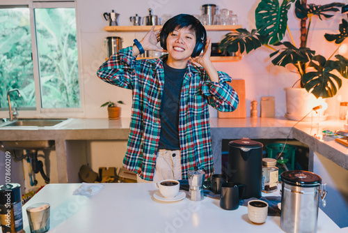 happy moment at home as an Asian woman enjoys her coffee, listening to music in the kitchen smiling in her plaid shirt and headphones, capturing a candid joyful portrait of everyday life.