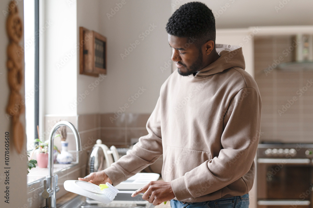Young man washing dishes in a cozy kitchen during the afternoon with natural light pouring in through the window Young man washing dishes in a cozy kitchen during the afternoon with natural light pouring in through the window