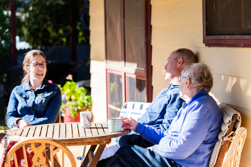 Three generations of family sitting around table on verandah in winter sunshine chatting together Three generations of family sitting around table on verandah in winter sunshine chatting together