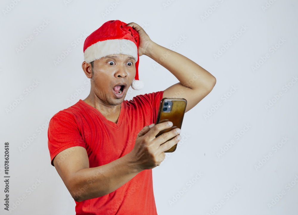 Shocked young Asian man in a Christmas hat looking at smartphone getting great news isolated over white background. celebration Christmas holiday and New Year 2025 concept.