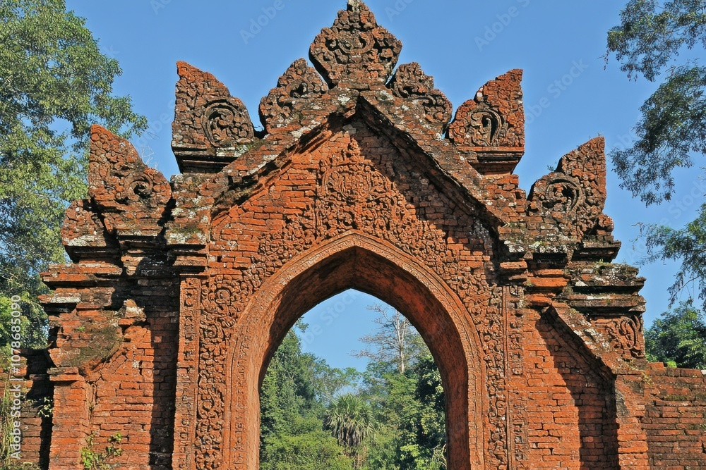 Naklejka premium Ancient red brick archway surrounded by lush greenery under a clear blue sky.