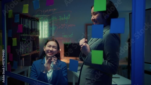 Two young businesswomen are discussing strategy in the office at night. They are using a glassboard with financial data and colorful sticky notes photo