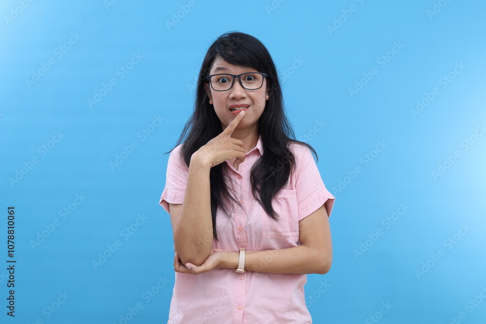 Photo of confused Asian girl bites lips and looks nervous at camera, holds hands partly crossed, dressed in casual shirt, stands against blue wall with blank space aside Photo of confused Asian girl bites lips and looks nervous at camera, holds hands partly crossed, dressed in casual shirt, stands against blue wall with blank space aside