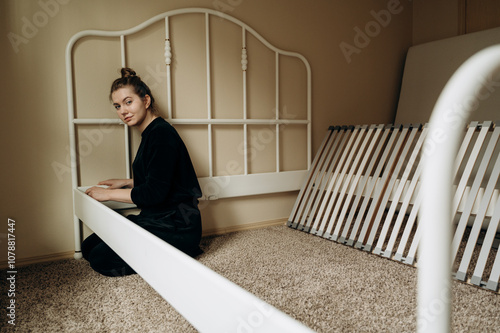 A handyman in a black suit assembles a metal bed by himself, sitting on a carpet. photo