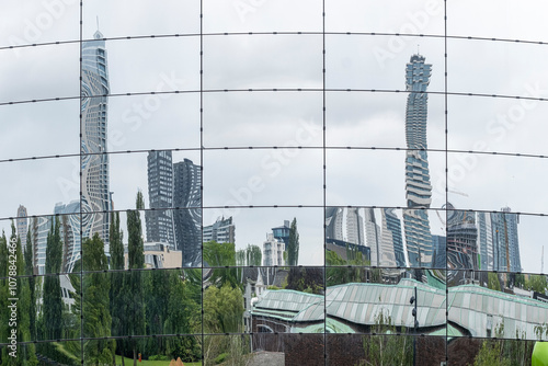 The reflective glass facade shows a distorted cityscape, merging buildings and nature into an abstract visual symphony, taken from across a lush green park. photo