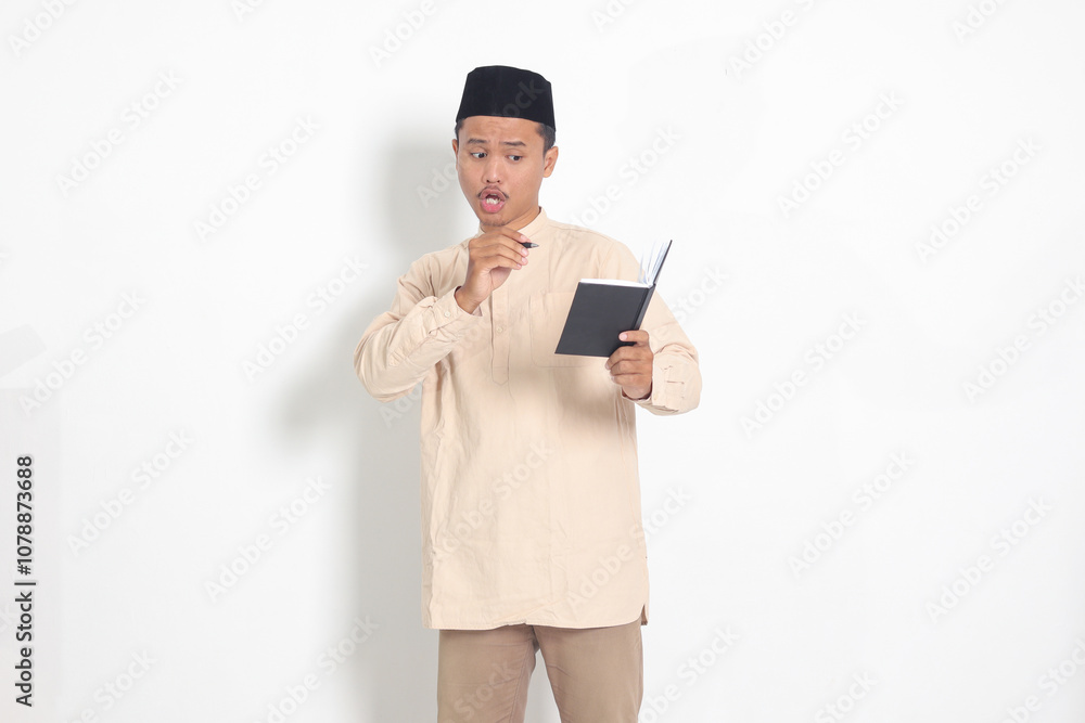 Portrait of excited muslim man in koko shirt with peci writing on his notebook. Happy Asian guy taking notes. Isolated image on white background Portrait of excited muslim man in koko shirt with peci writing on his notebook. Happy Asian guy taking notes. Isolated image on white background