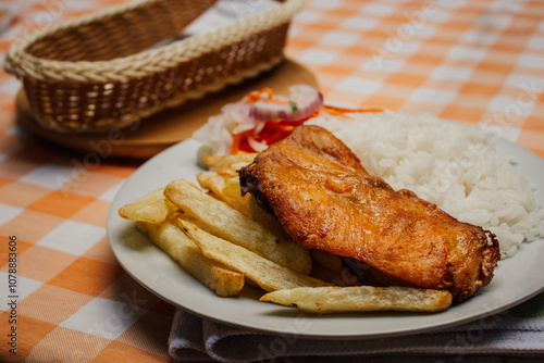 Pollo frito, acompañado de papas fritas y arroz blanco en plato blanco, sobre mesa a cuadritos anaranjado, con cubiertos a un lado photo