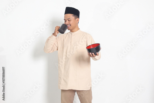 Portrait of excited Asian muslim man in koko shirt with skullcap holding a mug and empty bowl. Bowl template for food brand. Isolated image on white background photo