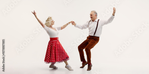 Cheerful elderly couple dancing together, woman in red polka dot skirt and man in suspenders isolated on white background. Concept of retro dance, hobby, aging, active lifestyle photo