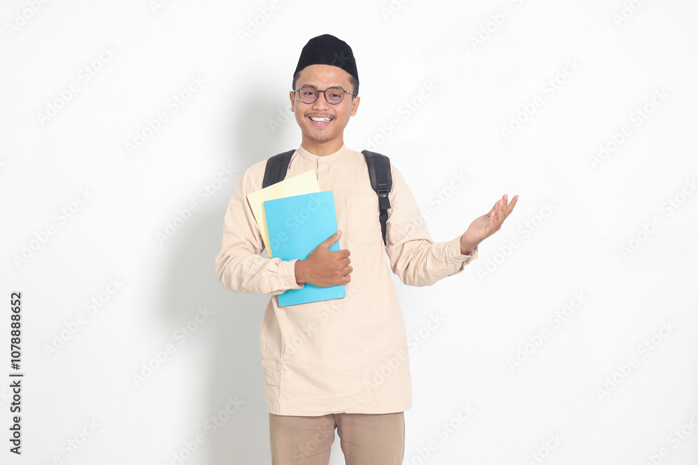 Portrait of excited student Asian muslim man in koko shirt with skullcap carrying backpack, holding school books, pointing to the side. Islamic education concept. Isolated image on white background Portrait of excited student Asian muslim man in koko shirt with skullcap carrying backpack, holding school books, pointing to the side. Islamic education concept. Isolated image on white background