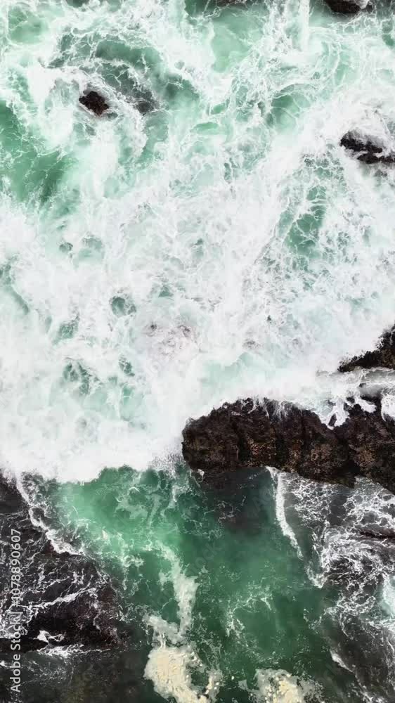 Aerial top view the ocean waves crash against the black rocks, creating white foam that contrasts with the dark green seawater. The water flow appears strong, breaking through the rough rock crevices. Aerial top view the ocean waves crash against the black rocks, creating white foam that contrasts with the dark green seawater. The water flow appears strong, breaking through the rough rock crevices.