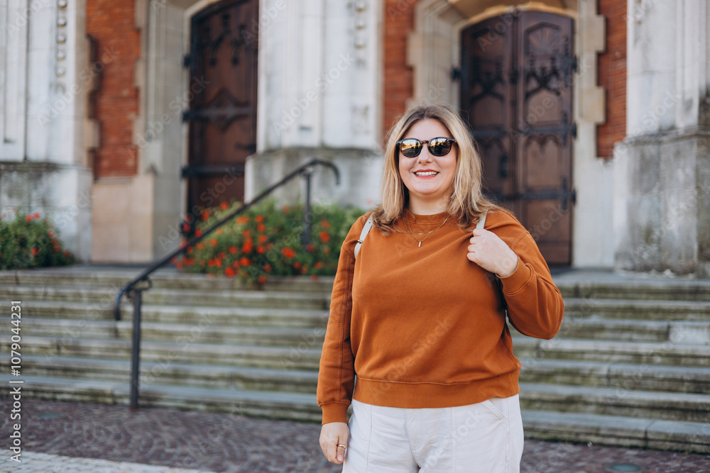 Fototapeta premium Happy Tourist girl walking in the city during vacation. Cheerful 30s woman in sunglasses traveling abroad in summer. Travel concept, old town square. Smiling model posing in the street