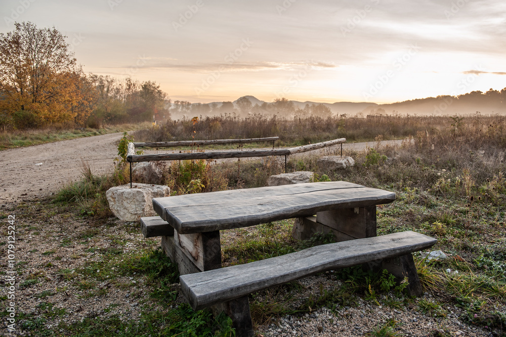 Grob gezimmerter Holztisch mit Sitzbänken in herbstlicher Landschaft mit Hügelkette und Nebel vor Sonnenaufgang Grob gezimmerter Holztisch mit Sitzbänken in herbstlicher Landschaft mit Hügelkette und Nebel vor Sonnenaufgang