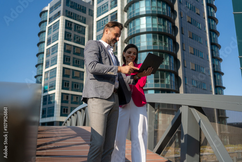 Smiling Latin Hispanic mature adult businesspeople using pc digital computer. Laughing caucasian colleagues, checking the company budget on their laptop in front of glass building exterior.	