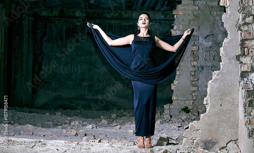 A beautiful young girl model stands in a long black dress amidst the rubble of a partially demolished building. photo