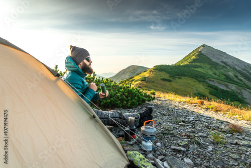 Stylish bearded man wearing down jacket sits beside his tent in front of a mountain peak grinding coffee photo