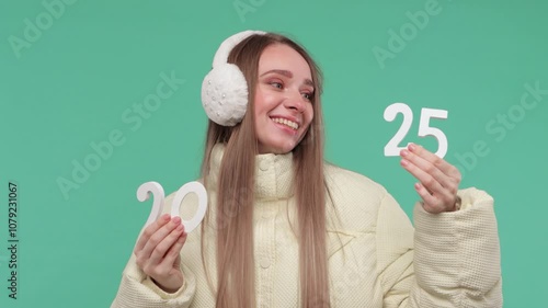 Wallpaper Mural A woman in ain warm yellow jacket and winter fur earmuffs on green background holds  numbers of the new year 2025. Torontodigital.ca
