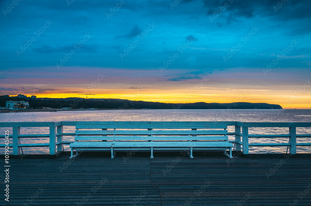 Fototapeta premium The inviting white bench stands on sopot pier at sunrise, offering a peaceful view of the colorful sky and tranquil baltic sea, creating a serene moment in the popular polish resort town