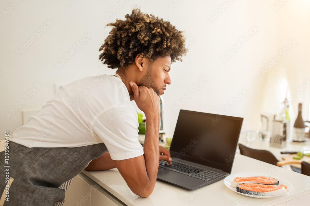 A young black man in an apron leans against a kitchen countertop, focused on his laptop as he looks up recipes. Several pieces of salmon sit on a plate beside him, indicating he's preparing a meal. A young black man in an apron leans against a kitchen countertop, focused on his laptop as he looks up recipes. Several pieces of salmon sit on a plate beside him, indicating he's preparing a meal.