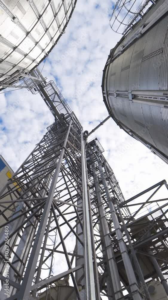 Metal construction of a modern agricultural factory. Industrial plant for storage and processing seeds. Grain elevators outdoors. View from below. Vertical video Metal construction of a modern agricultural factory. Industrial plant for storage and processing seeds. Grain elevators outdoors. View from below. Vertical video