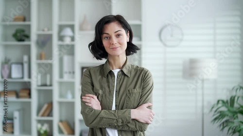 Serious young adult woman with dark hair stands confidently with crossed arms in modern apartment interior. Headshot of confident female in casual shirt posing looking at camera in living room at home