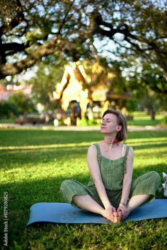 a young woman of European appearance sits in the lotus position on a blue rug in a city park. Yoga in the park 