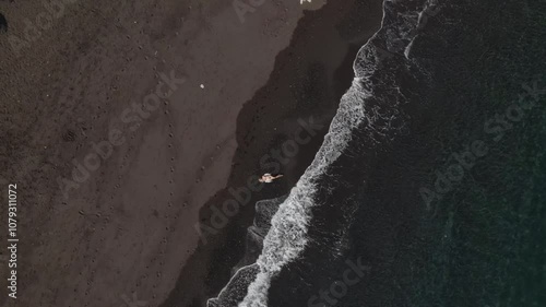 Drone Approaching Woman with Light Brown Hair in White Dress Relaxing on a Black Sand Beach by the Ocean Waves.