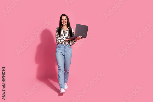 Young woman with brunette hair in casual jeans and a striped shirt holds a laptop against a vibrant pink background, smiles confidently