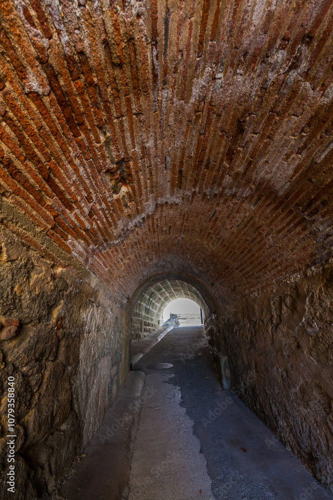 An arched brick tunnel with textured, aged walls leading towards an exit illuminated by bright daylight An arched brick tunnel with textured, aged walls leading towards an exit illuminated by bright daylight