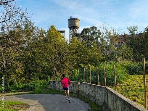 Rome, Italy - November 13, 2024: A runner jogs along the Tevere cycle path at Lungotevere di Pietra Papa, surrounded by lush greenery and urban graffiti in the Marconi district of Rome.
 photo