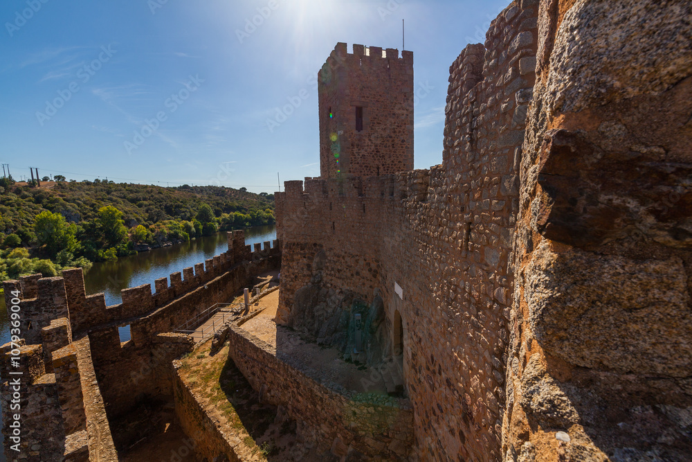 The rugged stone walls of the castle stand tall, overlooking a serene river that flows alongside the fortress The rugged stone walls of the castle stand tall, overlooking a serene river that flows alongside the fortress